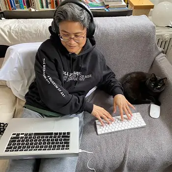 Kamomi working on her couch while her cat stares at the camera